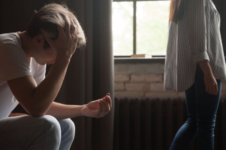 A man sits on the bed with his hands on his head and a woman walks away