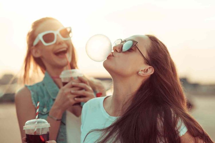 Two young ladies laughing in the sunset with ice coffee and one blowing a bubble