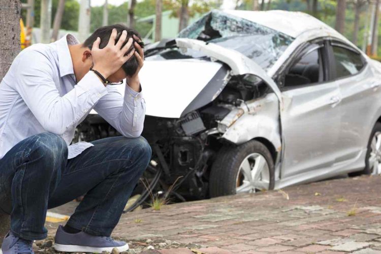 A man kneeling by a tree with his head in his hands and a crashed car in the background