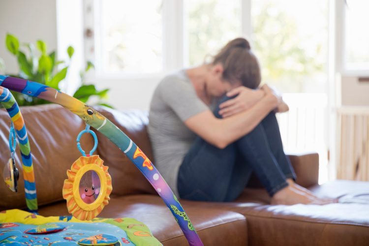 A sad woman hugging her knees on the couch with a baby toy in the foreground